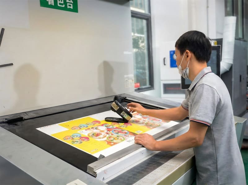 Production staff performing printing color calibration, checking color accuracy on a press sheet using professional measurement tools.